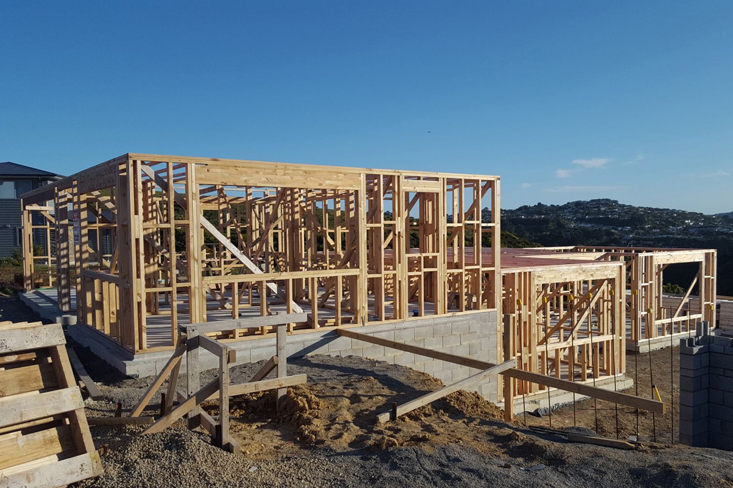 Wooden framework of a house under construction on a concrete foundation, hillside setting, clear blue sky.