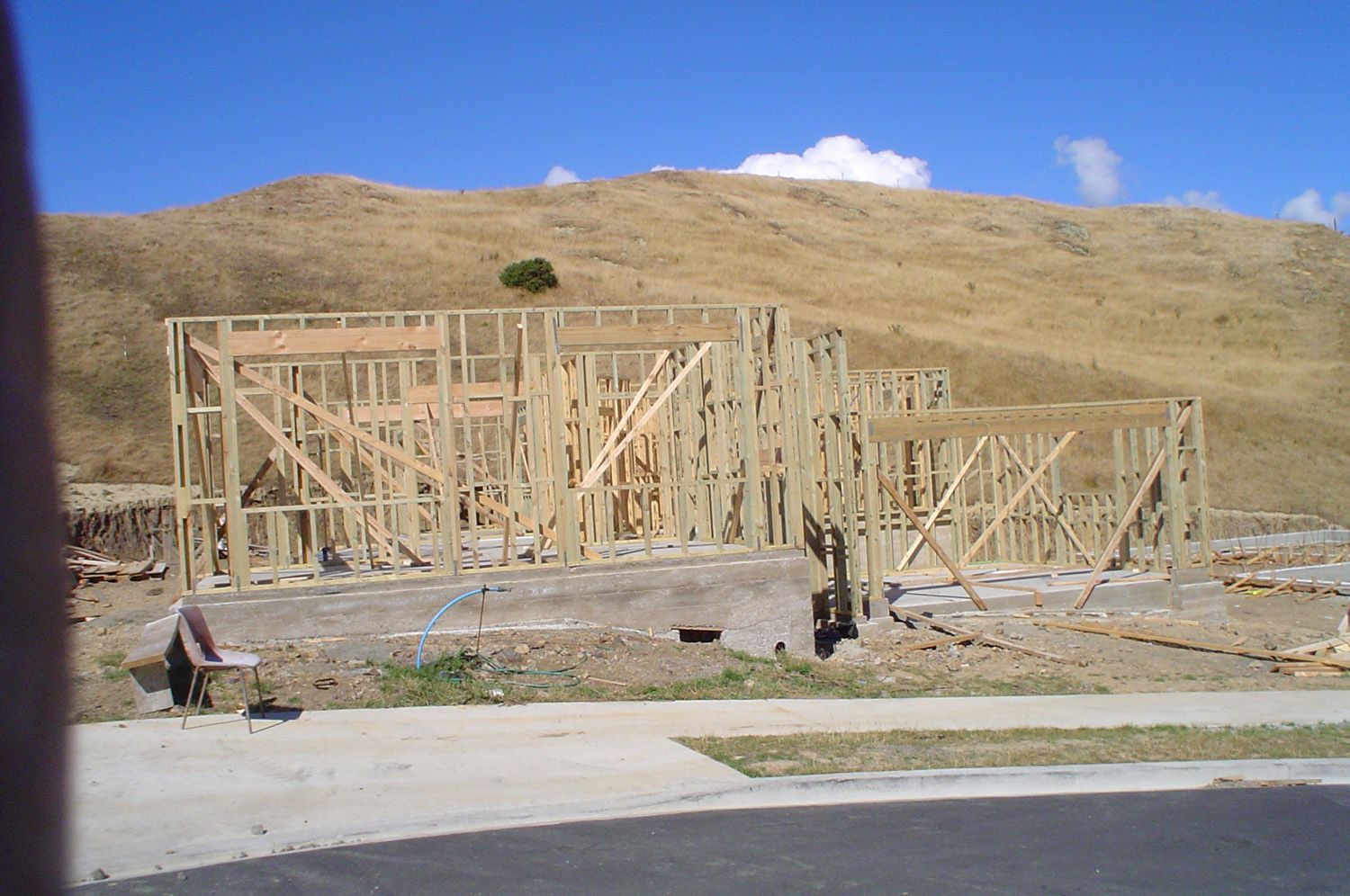 Wooden frame of a house under construction on a concrete foundation, with a dry, brown hillside in the background.
