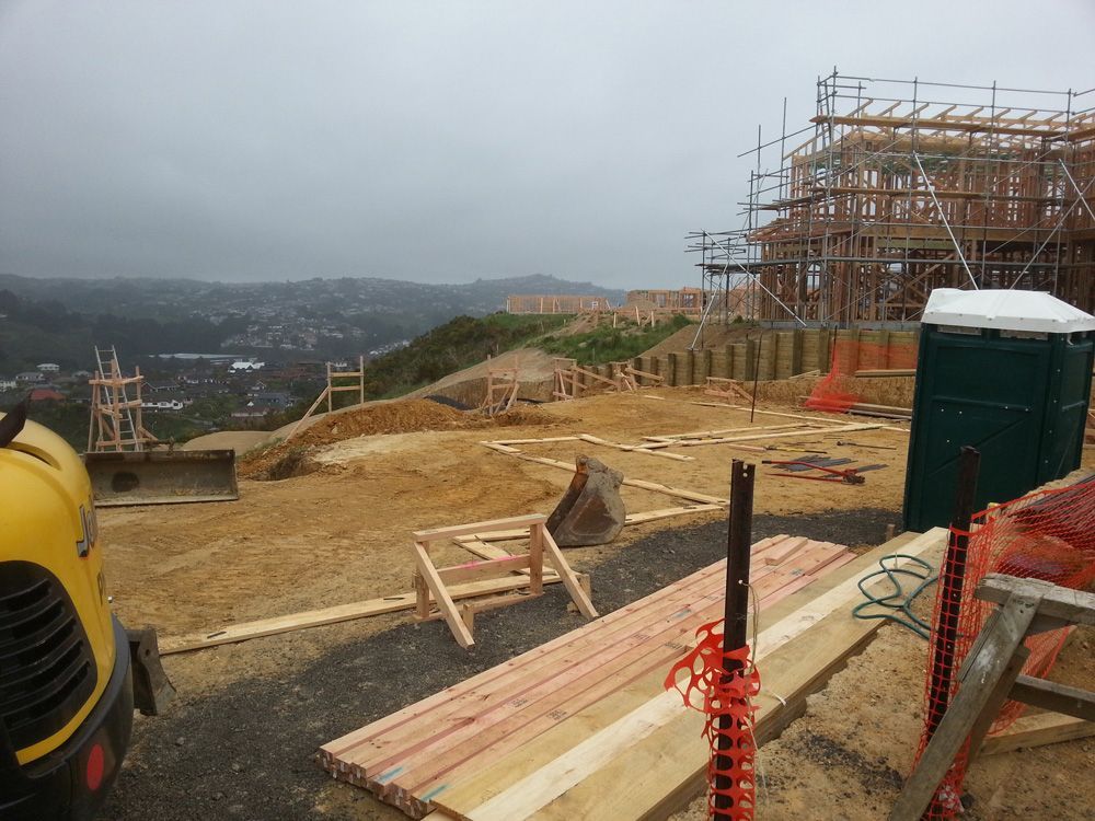 Construction site on a hillside, wood framing of a house, soil, excavator, portable toilet, overcast sky.