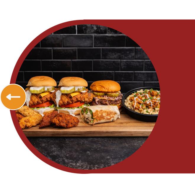 Assorted burgers and fried chicken on a wooden board against a dark brick wall, with a side dish.