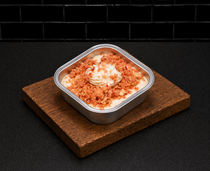 Square dessert in a metal dish topped with cream, resting on a wooden board against a dark background
