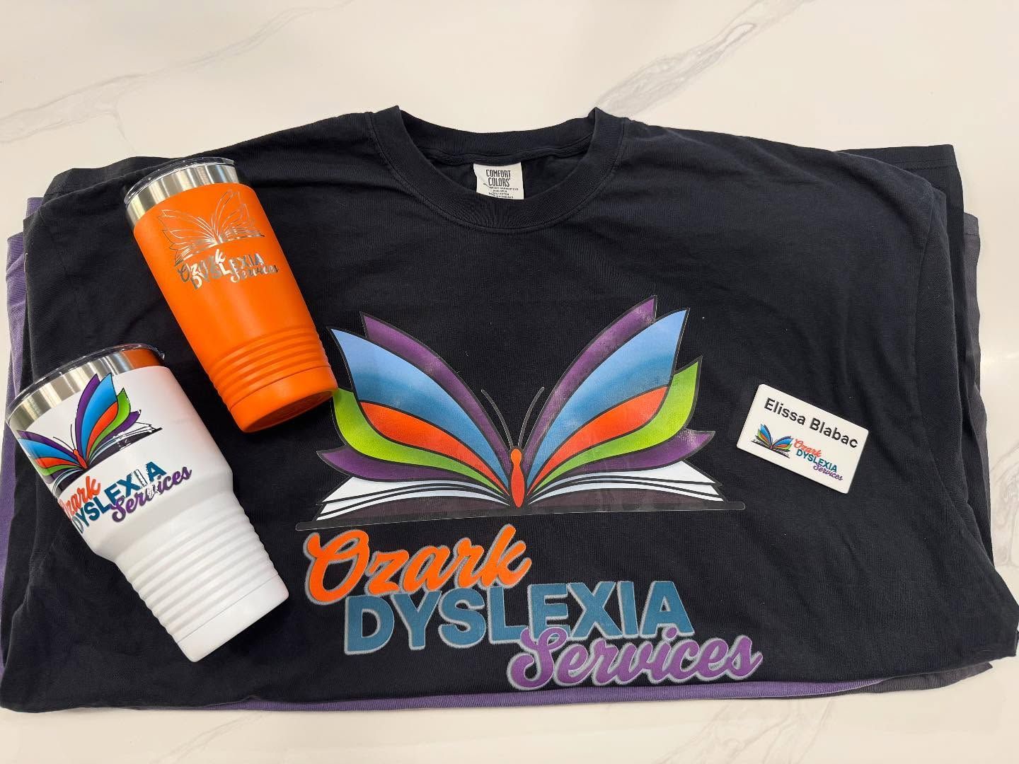 Black Ozark Dyslexia Services T-shirt with colorful feather logo and two branded cups on top