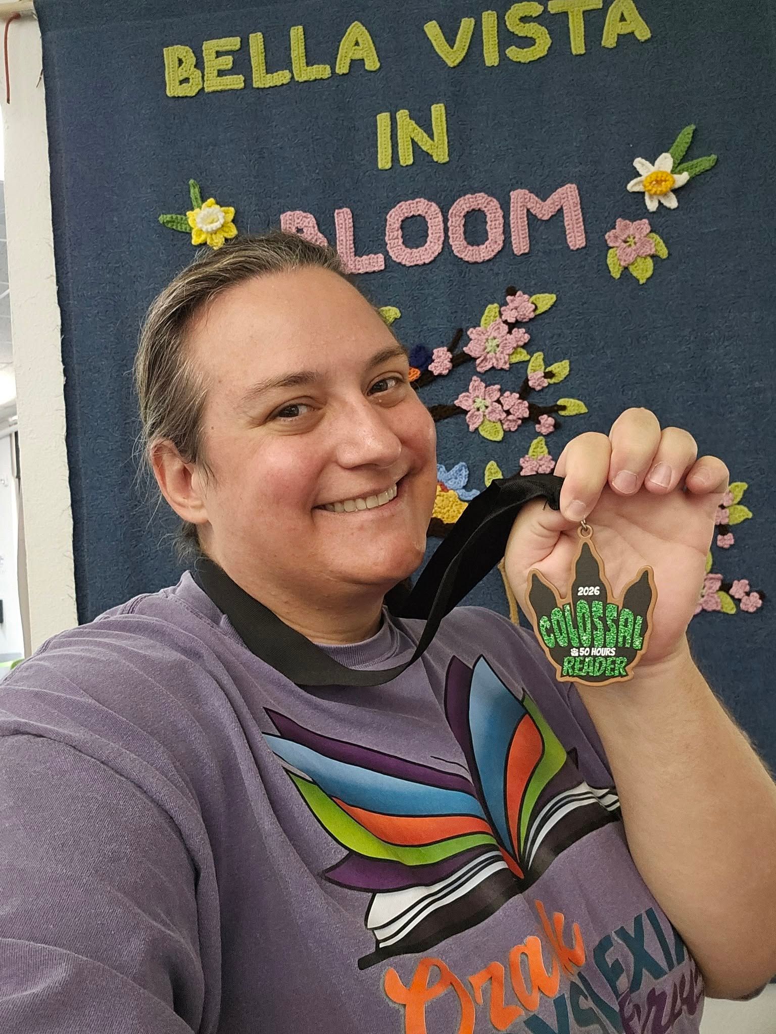 Smiling person in front of a Bella Vista in Bloom sign, holding a green flower-shaped keychain