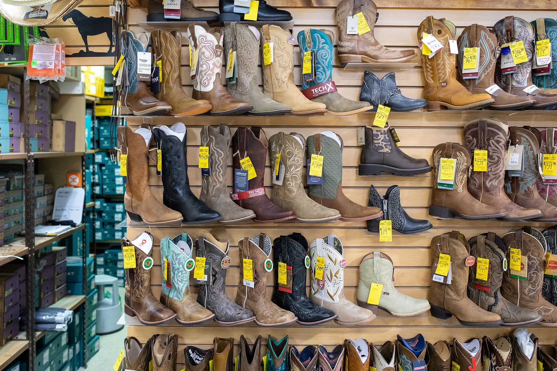 Wall of boots on shelves in a shop, with yellow price tags and stacked boxes on the left.