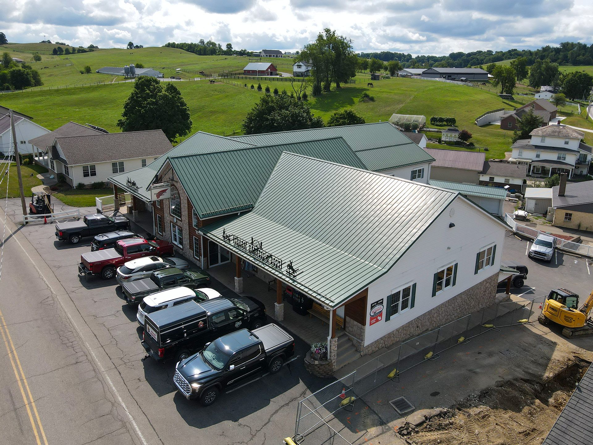 Aerial view of a white building with green roof, parked cars, and grassy fields in the background.