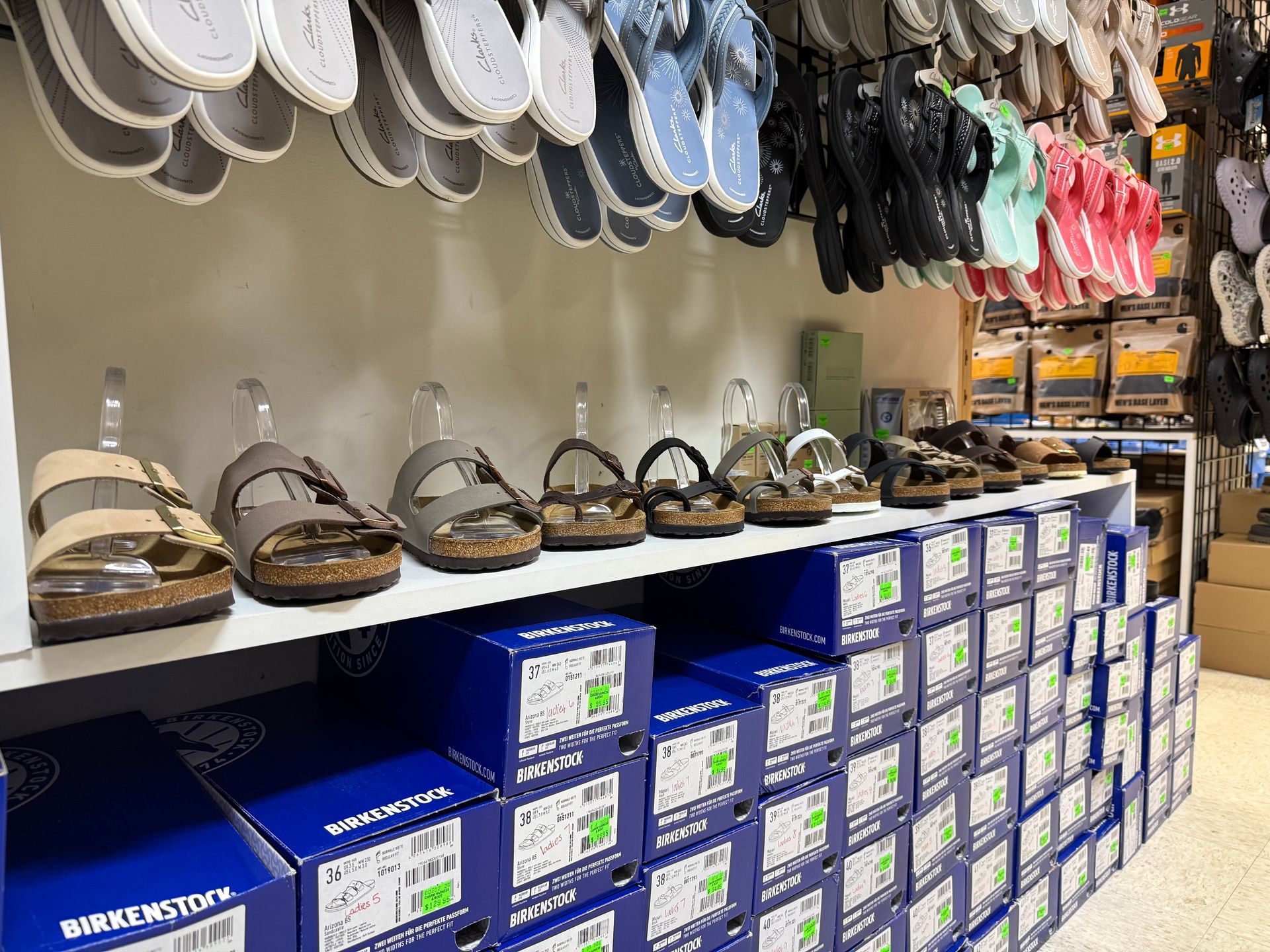 Shoe store aisle with sandals on display and stacked boxed footwear below.