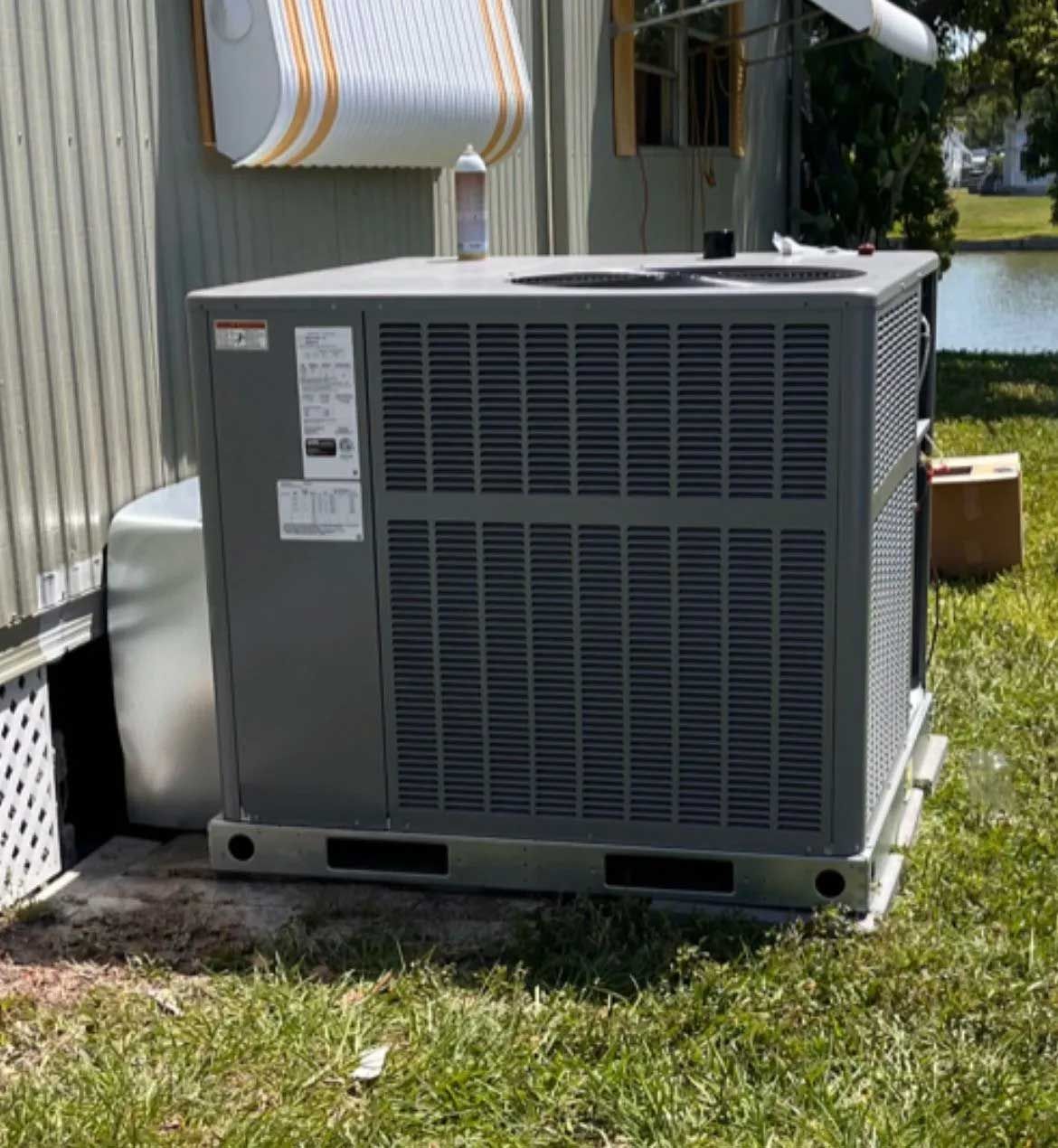 Outdoor HVAC unit beside a house on grass near a lake, with a utility cover overhead.