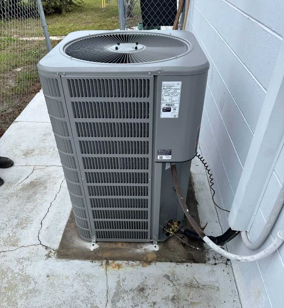 Outdoor air conditioner unit beside a white wall on a concrete pad, with visible hoses and wiring.