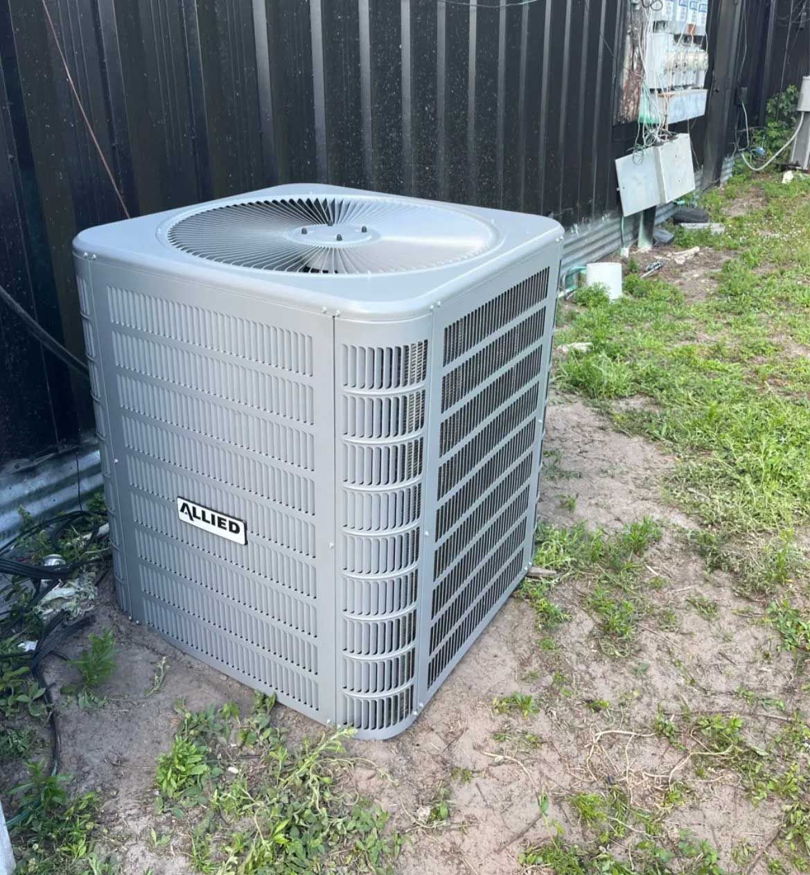 Gray outdoor air conditioner unit beside a black fence on a grassy patch