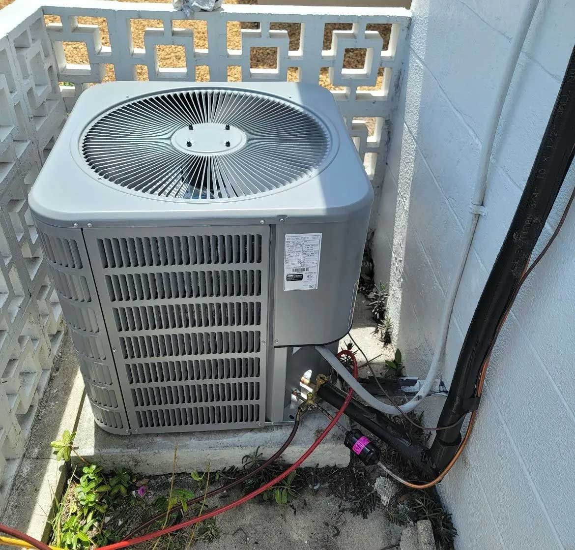 Outdoor air conditioner unit beside a wall, with red and black hoses on the ground.