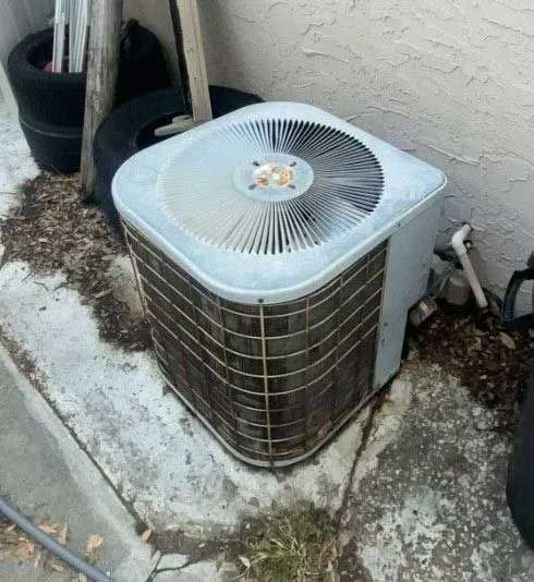 Outdoor air conditioner unit beside a house wall on a concrete pad with weeds and debris nearby