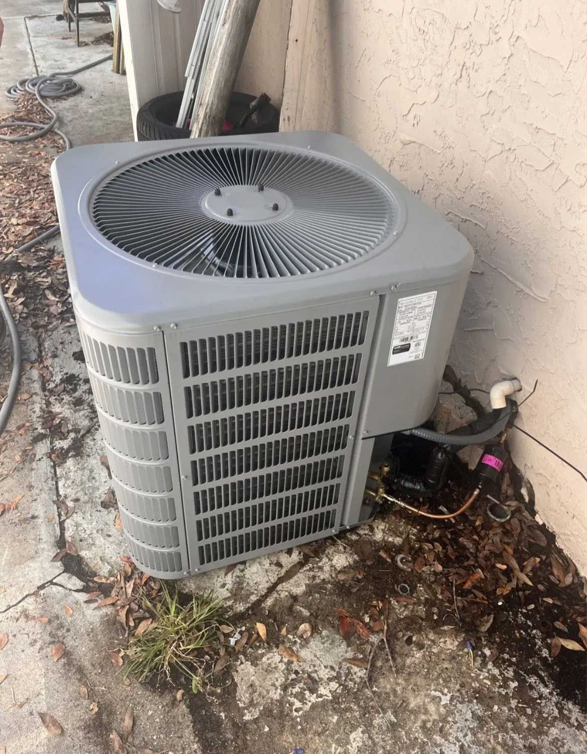 Outdoor air conditioner unit beside a stucco wall with debris on the ground and exposed wiring nearby
