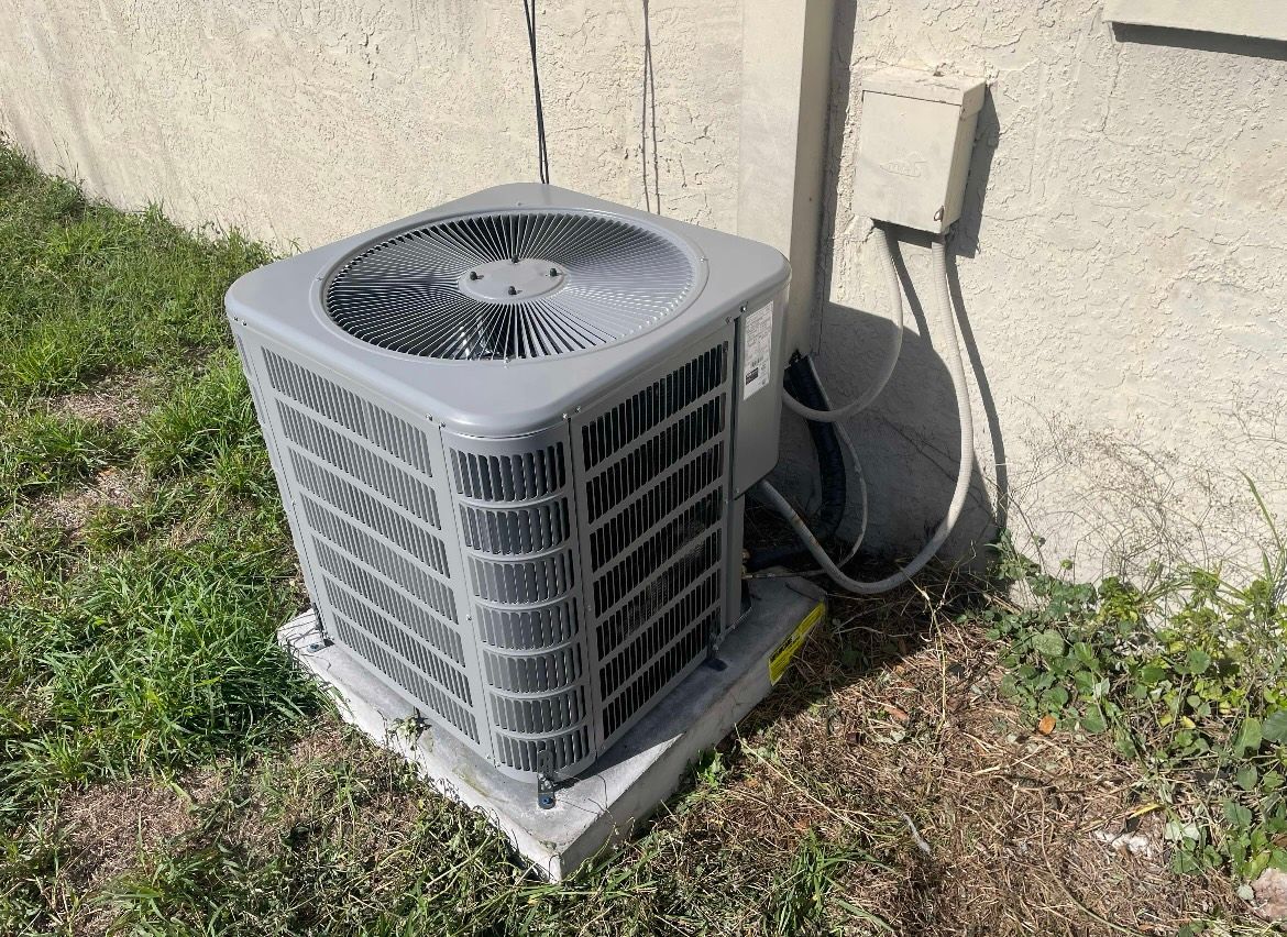 Outdoor air conditioner unit beside a stucco wall on a concrete pad, with grass nearby