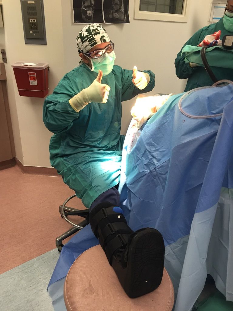 Surgeon in scrubs gives thumbs up, next to a patient's leg with a medical boot in an operating room.