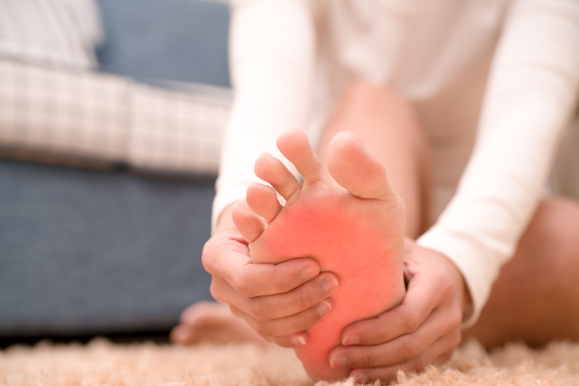 Person holding foot, showing red area indicating pain. Sitting on carpet indoors.