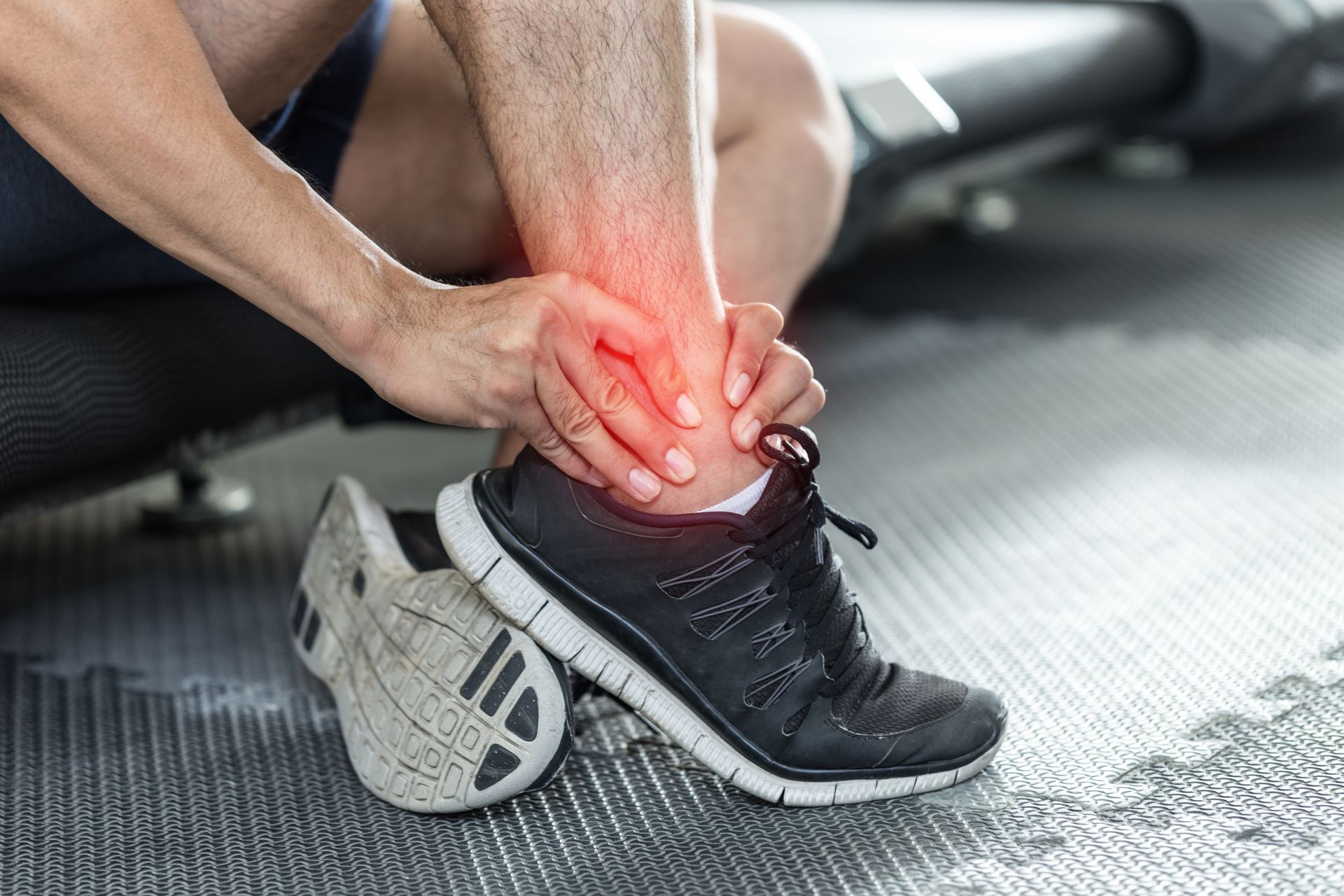 Person holding ankle, showing red pain area, sitting near treadmill and wearing sneaker.