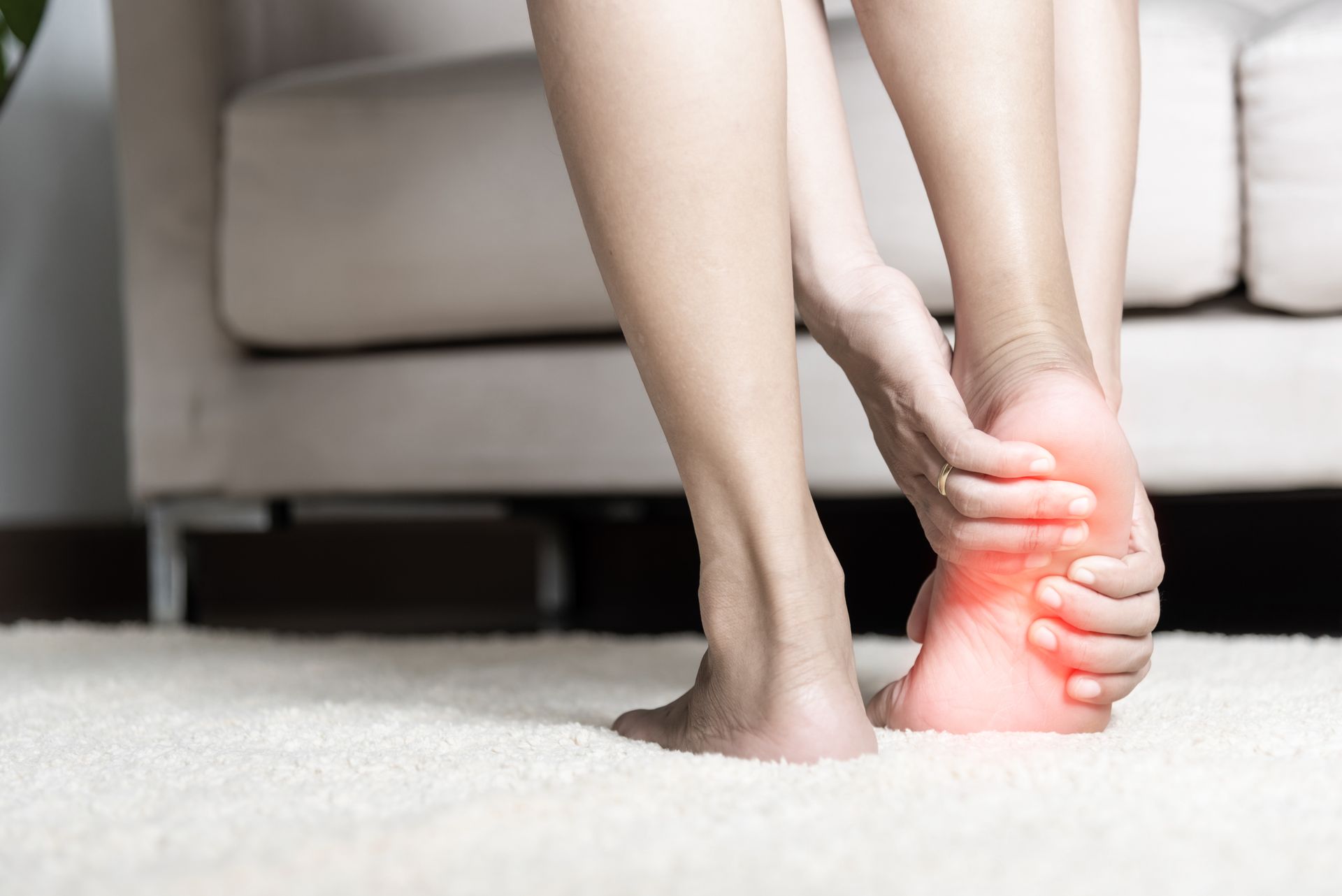 Woman holding foot, indicating pain; red glow on heel; indoors on a white rug.
