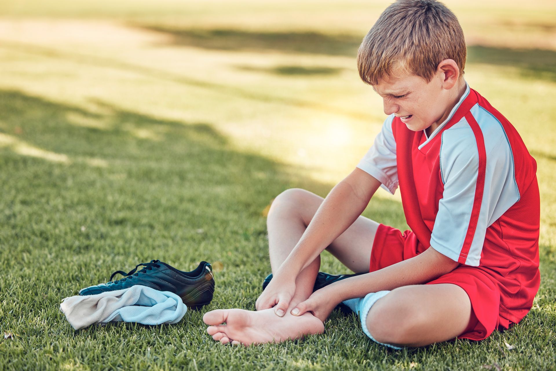 Boy sitting on grass, holding ankle, appearing pained. Soccer shoe and sock nearby.