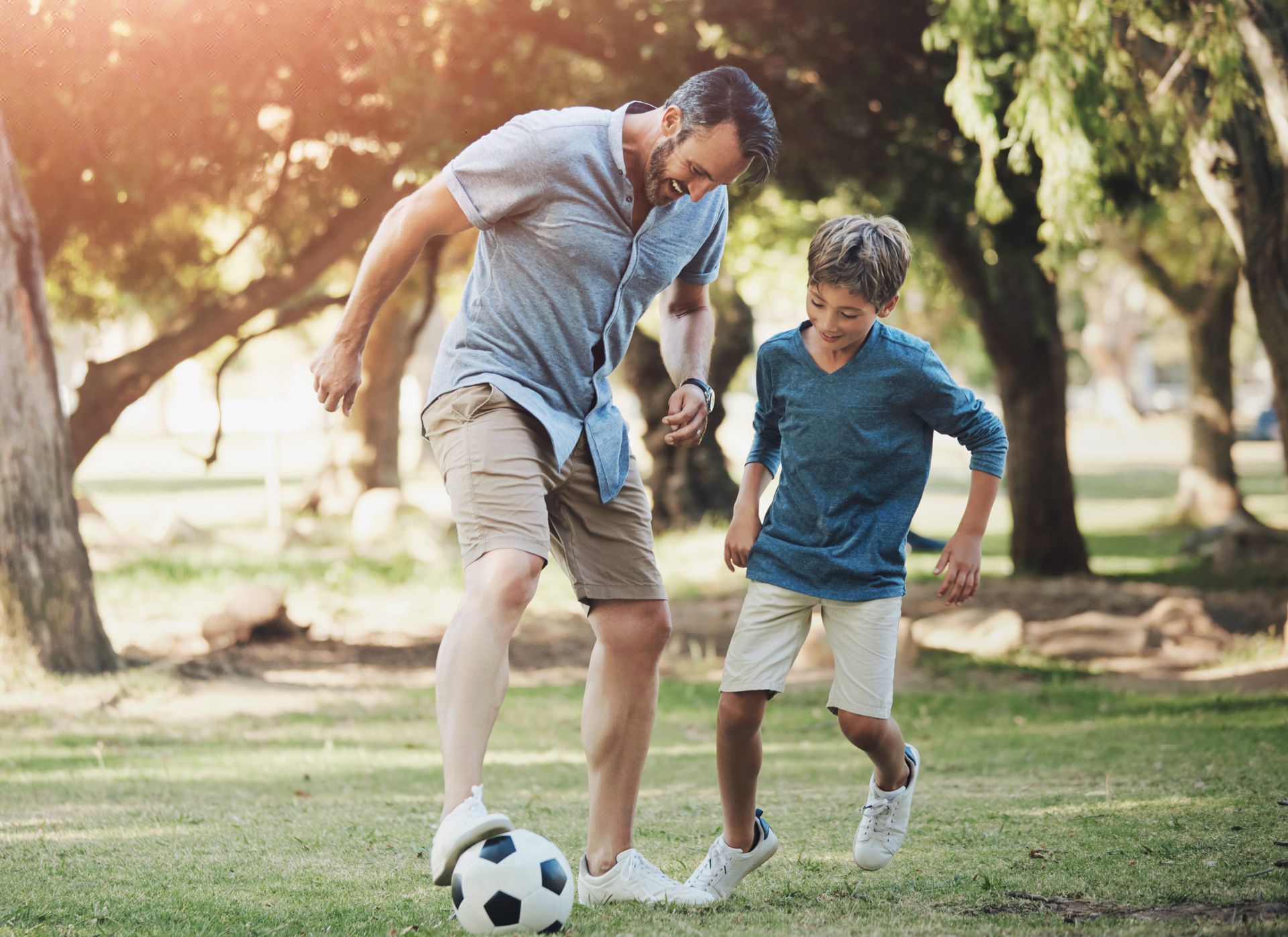 Father and son playing soccer in a park; sunny day, father kicks the ball, son watches excitedly.