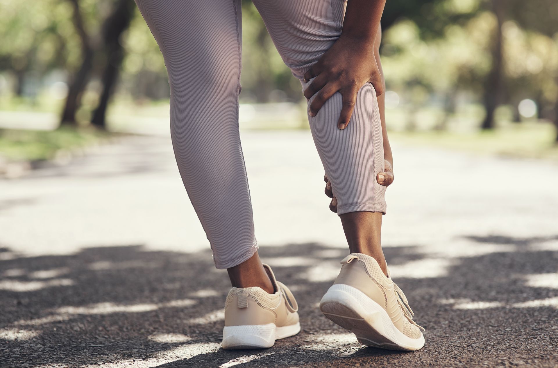 Person holding their calf in pain while standing on a path, wearing light-colored shoes and leggings.