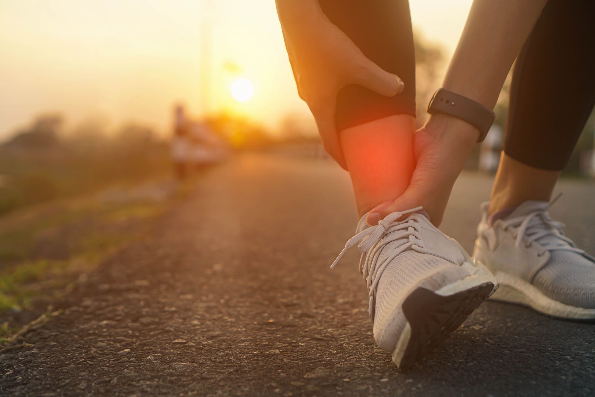 Person holding their ankle, indicating pain, while running outdoors at sunrise.