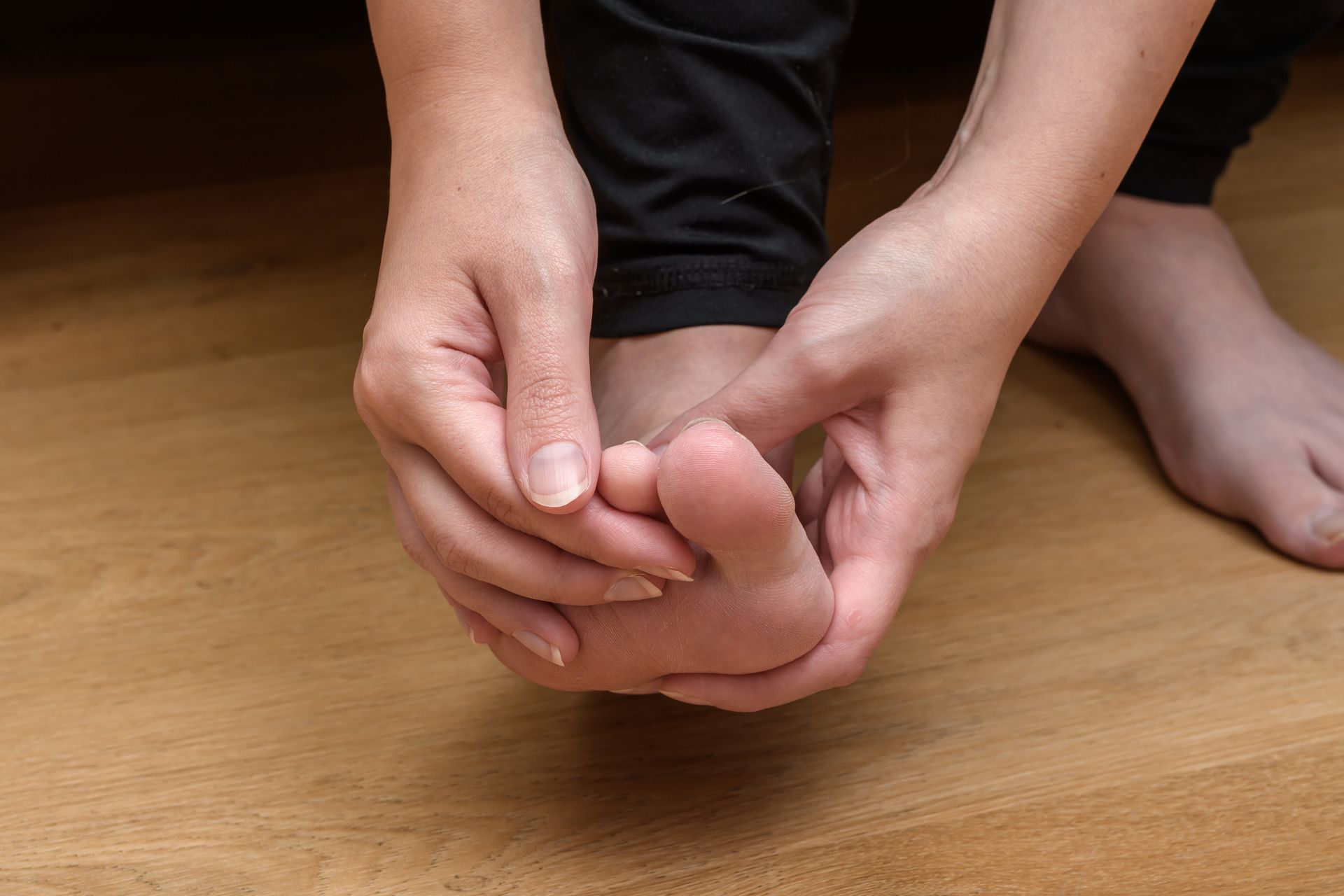 Person holding foot, examining it with hands, on a wooden floor, possibly experiencing pain.