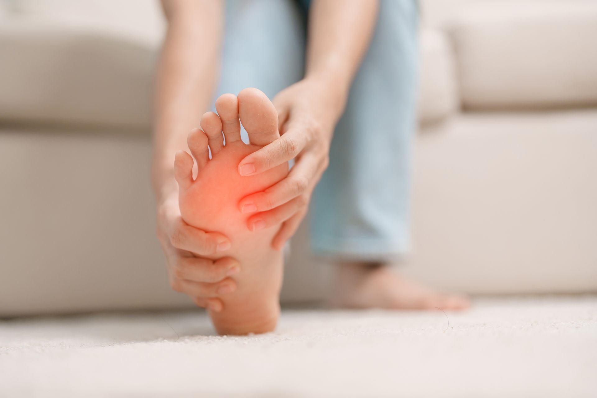 Person holding foot, pointing to a red area, indicating pain. Indoors on a carpet.