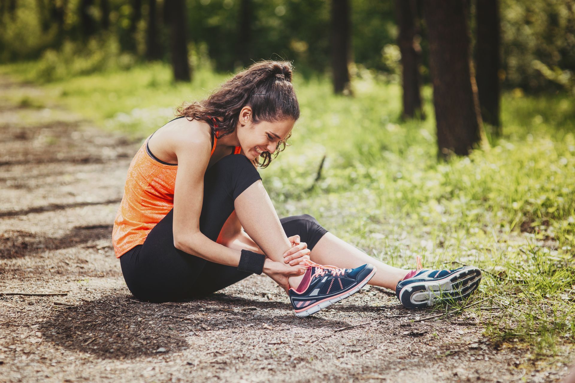 Woman sitting on path in pain, holding ankle, wearing athletic clothes, outdoors.