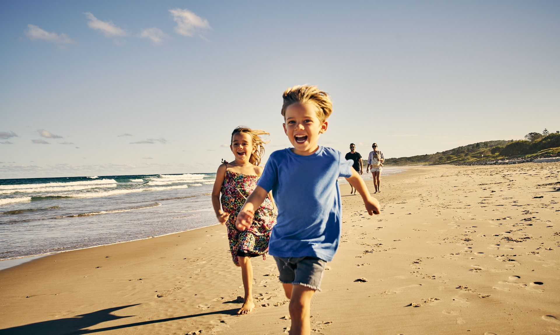 Two children running on a beach, smiling with ocean and family in the background.