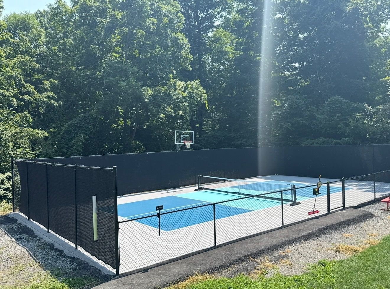 Outdoor fenced pickleball court with blue playing surface and basketball hoop, surrounded by trees.