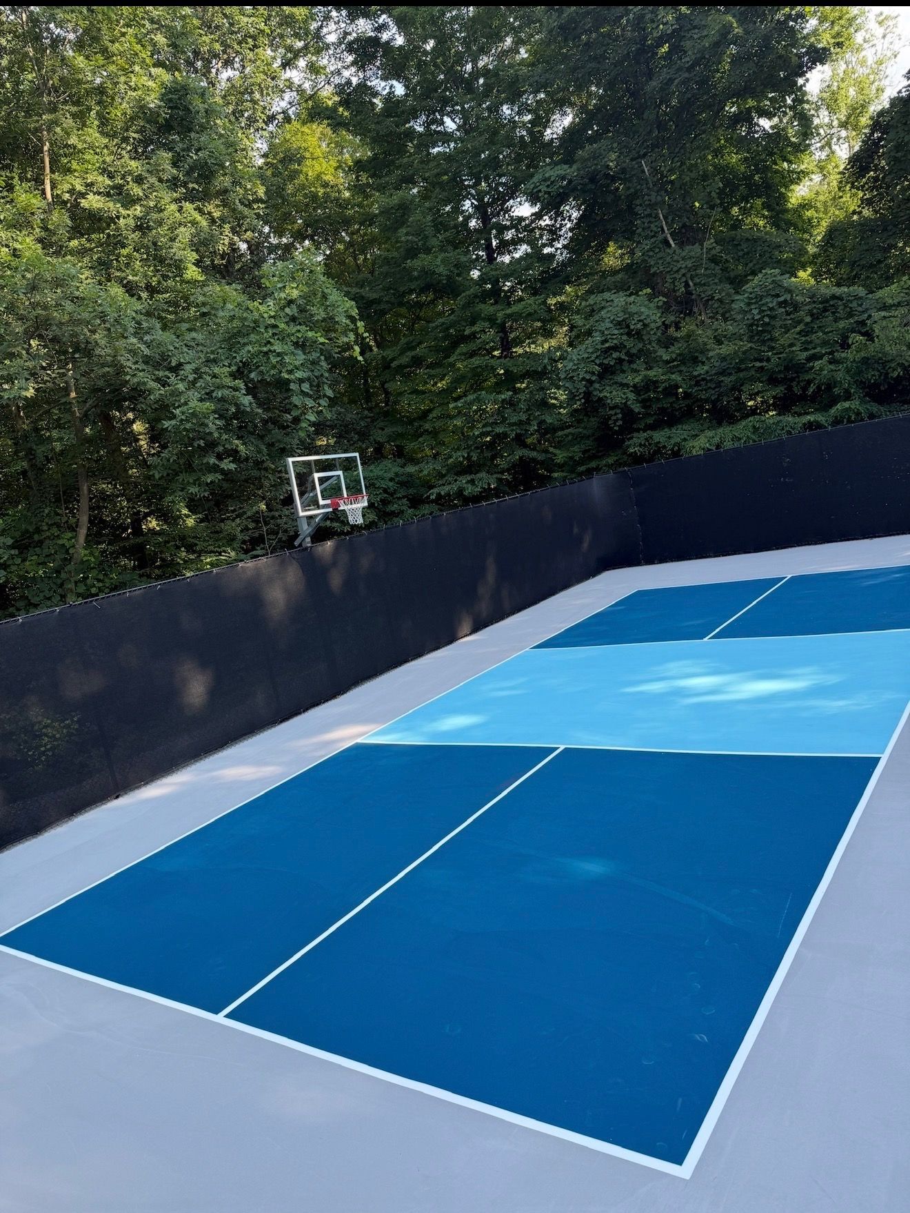 Blue tennis court surrounded by black fencing and trees, with a basketball hoop in the background
