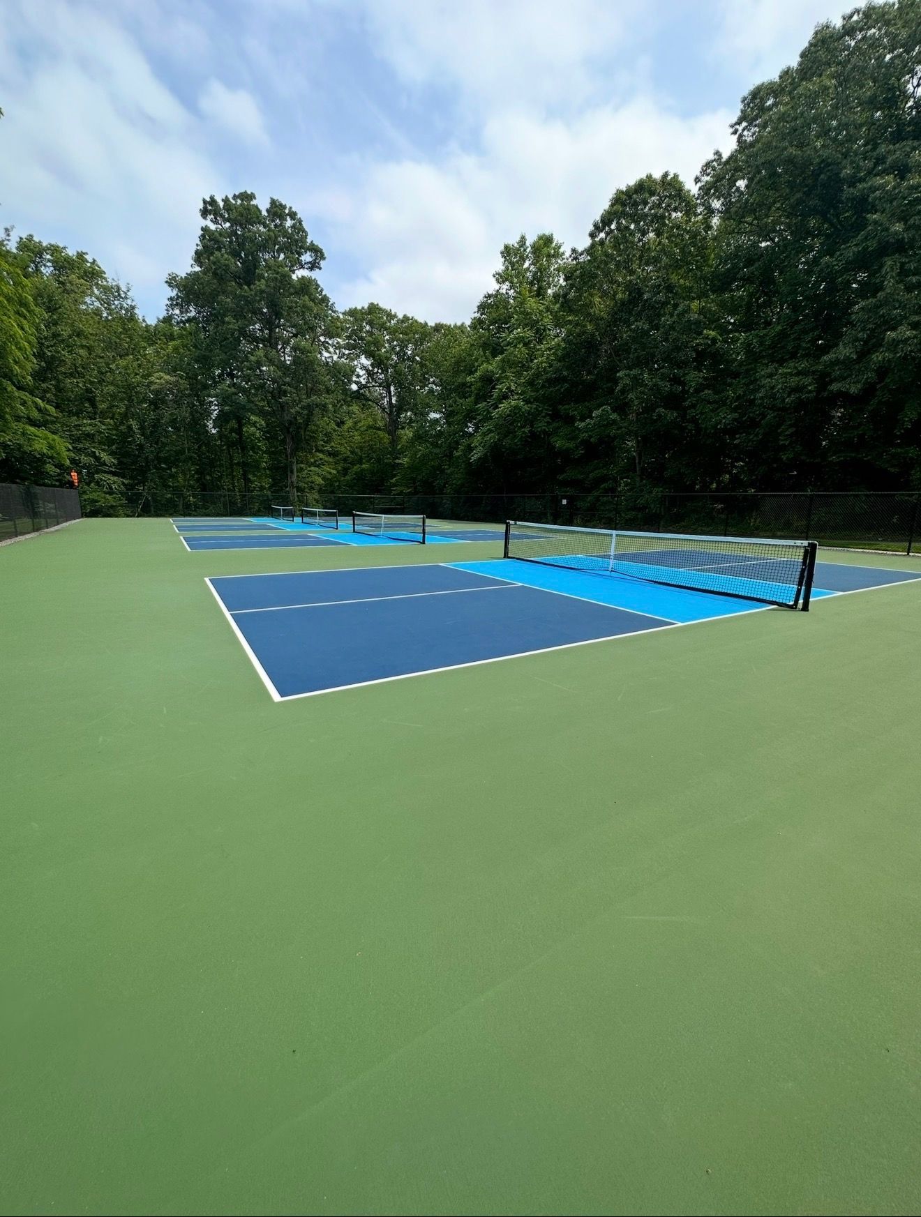 Empty outdoor tennis court with blue playing surface and green surroundings, bordered by trees.