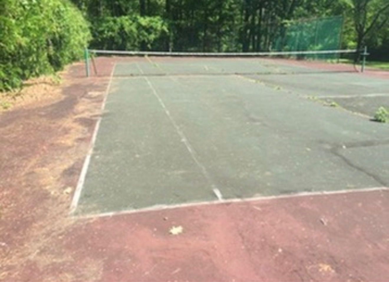 Empty outdoor tennis court with faded green surface, red borders, and trees around it.