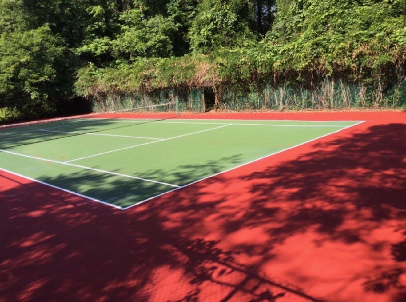 Outdoor tennis court with green playing surface and red perimeter, shaded by trees in sunlight