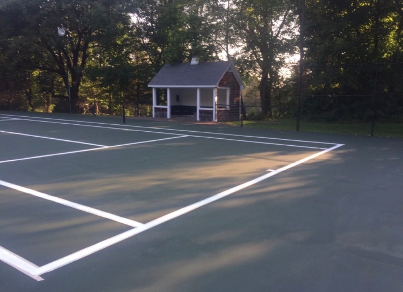 Empty outdoor tennis court with white lines and a small pavilion in the background at dusk