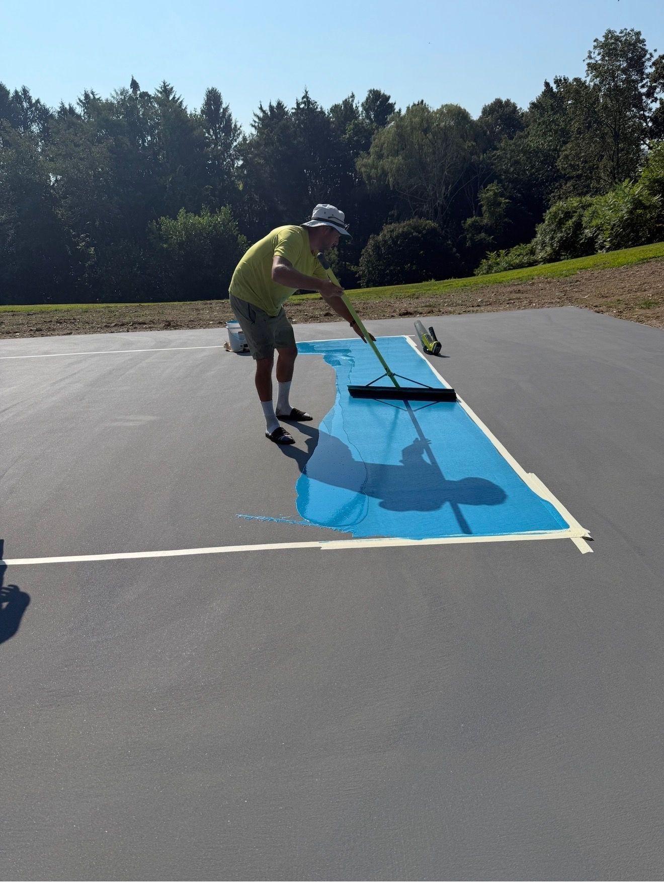Person painting blue lines on a paved court with a roller under bright sunlight