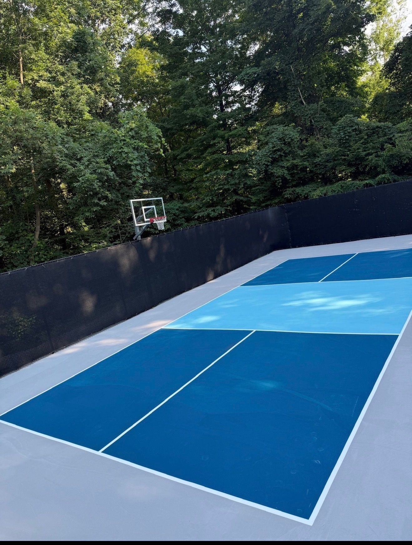 Outdoor blue-and-gray sports court beside a wooded area, with a hoop at the far end