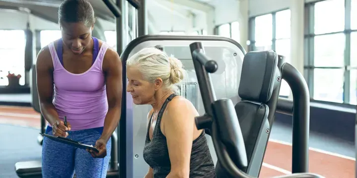 Woman in gym clothes talks to another woman at a workout machine, both in a gym.