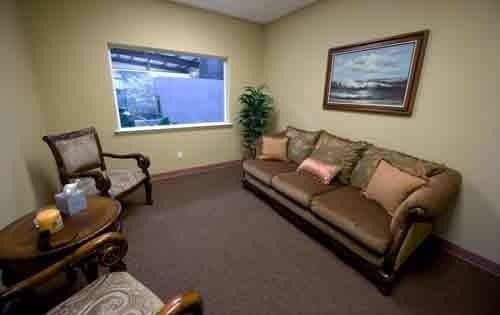 Small waiting room with beige sofa, chair, side table, window, and framed wall art.
