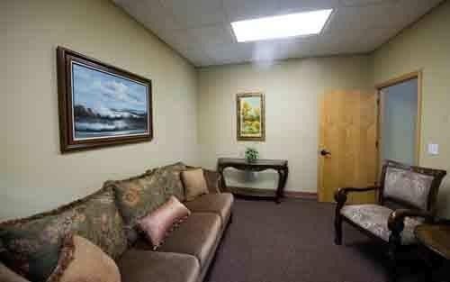 Small waiting room with a brown couch, two chairs, framed art, and a closed wooden door.