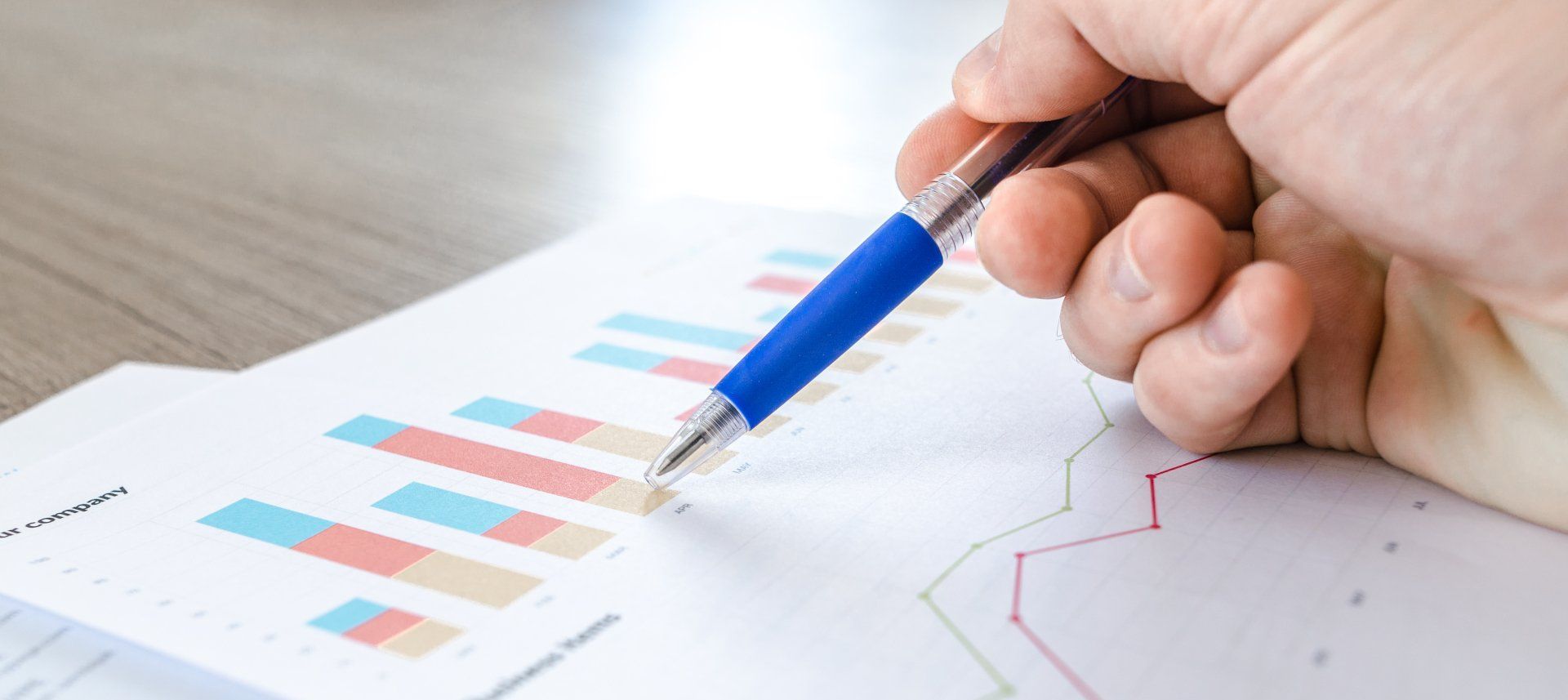 A hand holding a blue pen points to a bar graph and a line graph on a white paper.