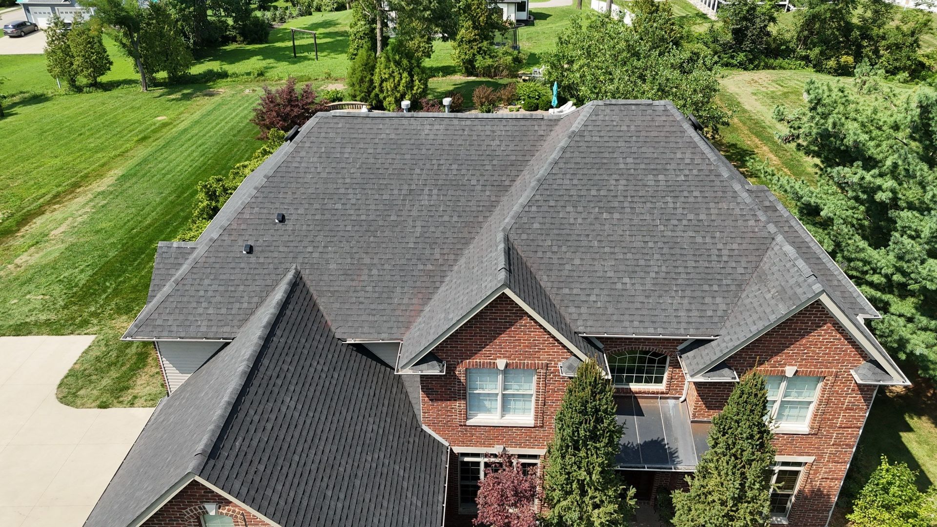 Aerial view of a large house with dark gray roof and brick exterior, surrounded by green lawn and trees