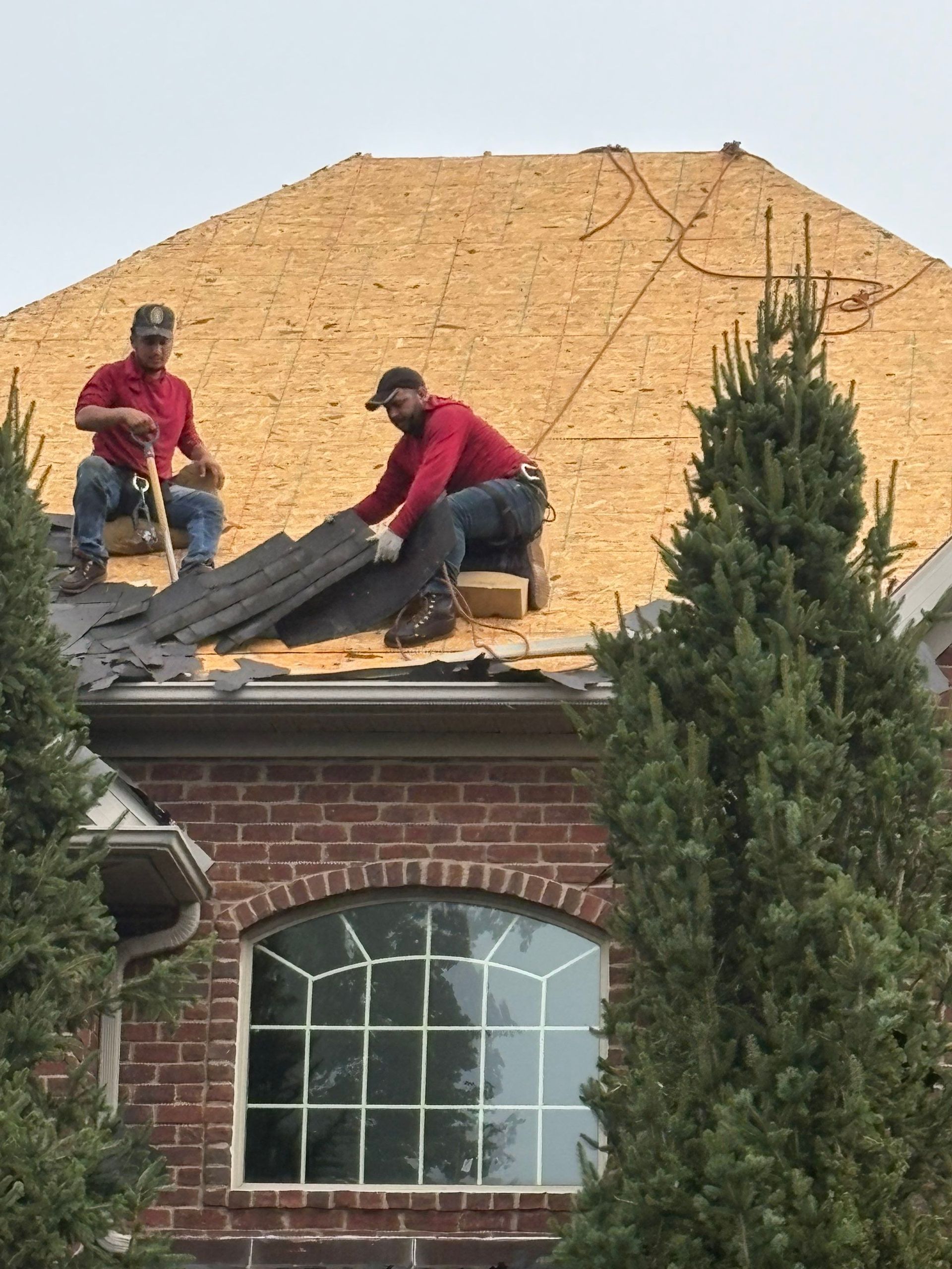 Two workers on a roof installing black shingles on a house with a brick facade and arched window.