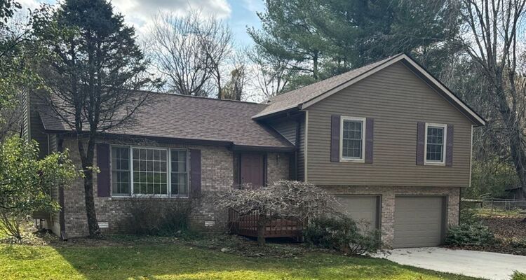 Suburban house with brown siding, attached garage, front porch, and trees in the yard.