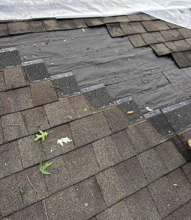 Damaged shingle roof with exposed underlayment and scattered leaves, viewed from above