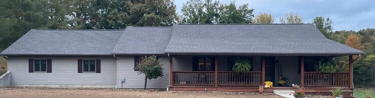 Single-story gray house with a dark roof, red brick porch, and trees in the background