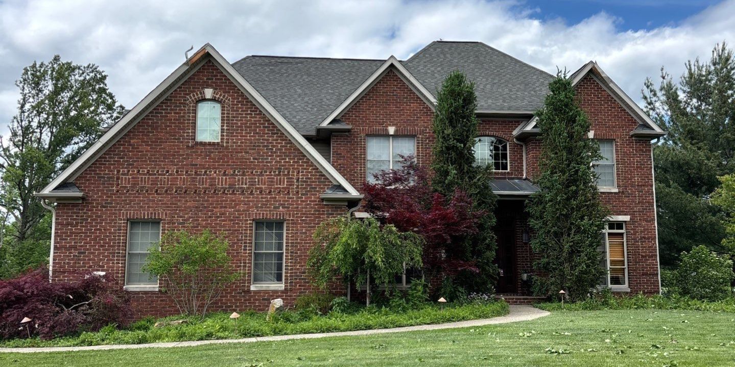 Red brick two-story house with gray roof, small windows, and overgrown front yard under a cloudy sky