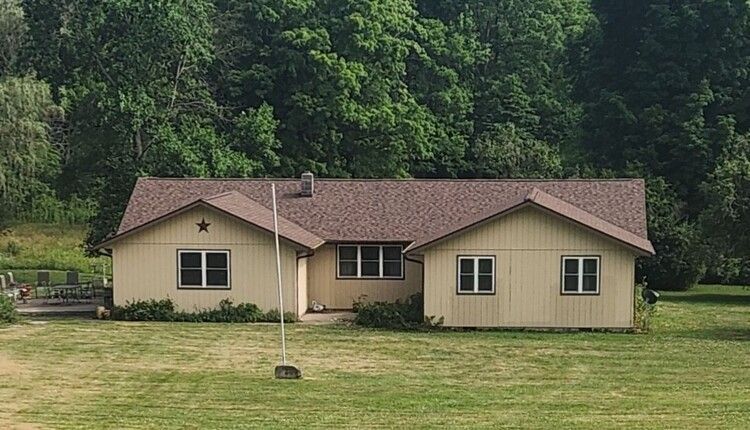 Small beige house with brown roof in a grassy yard, backed by dense green trees.