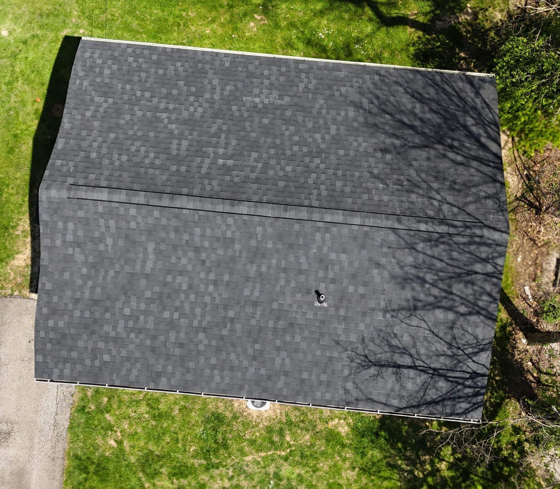 Aerial view of a gray shingled house roof beside green grass and tree shadows.