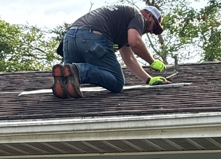 Worker kneeling on a shingled roof, using green gloves to repair the shingles.